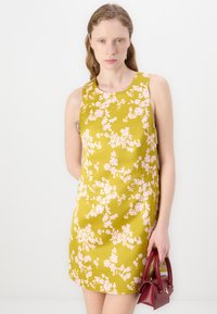 Rochie florală în galben muștar cu flori roz, design fără mâneci și decolteu rotund. Asortată cu o poșetă mică burgundă.