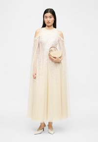 Donna in un abito lungo color crema con spalle scoperte e decorazioni scintillanti, che tiene in mano una piccola borsa beige e indossa tacchi a punta.