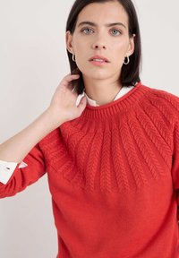 Röd stickad tröja med en texturerad kabelstickad design på den övre delen och en slät nedre del. Ribbad krage och ärmslut.