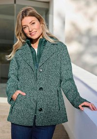 Lächelnde Frau mit blonden Haaren, die eine grüne gemusterte, knöpfbare Jacke über einer gesteppte, mit Reißverschluss versehenen Schicht trägt, steht im Freien neben einer weißen Wand.