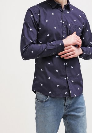 Muž, který má na sobě tmavě modrou košili na knoflíky s bílými motivy ptáků a světle modré džíny, stojí se sepjatýma rukama v pase.