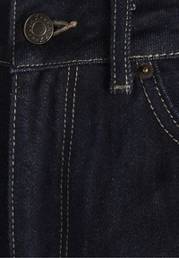 Tmavě modrá denimová látka zblízka, s měděným knoflíkem a kontrastním žlutým stehem podél pasu a bočního švu.