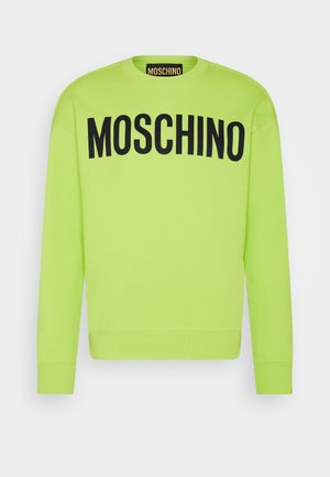 Henkilö pukeutunut neonkeltainen Moschino-huppariin ja mustiin shortseihin, istuu jalka kohotettuna näyttäen sukkia, joissa on sumeaa valkoista tekstiä.
