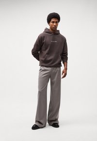 Bruine hoodie met een voorzak en witte tekst, gecombineerd met lichtgrijze wijdvallende broeken en zwarte schoenen. Gladde texturen, casual stijl.