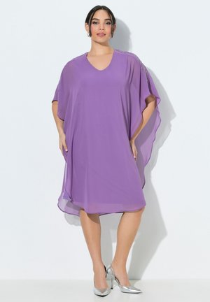 Frau trägt ein lockeres, knieumspielendes, lavendelfarbenes Kleid mit verzierten Schultern und silbernen High Heels, steht vor einem weißen Hintergrund.