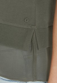 Grøn bomuldsblanding top med ribbet kant, sideskilt detaljer og en lille logo knap accent bagpå. Blød tekstur, afslappet pasform.