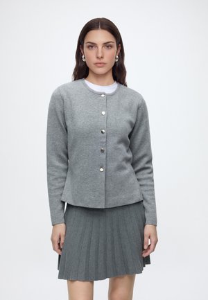 Femme aux longs cheveux bruns portant un cardigan gris boutonné par-dessus un haut blanc et une jupe plissée grise, debout sur fond blanc.