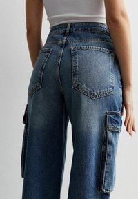 Pantalones cargo de mezclilla azul de cintura alta con bolsillos frontales y traseros, patas anchas y una textura desgastada. Bolsillos laterales ajustables acoplados.