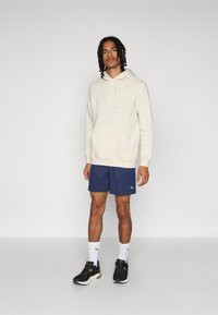 Haut à capuche beige clair avec une poche frontale et un logo PUMA, shorts navy, baskets noires et chaussettes blanches avec logo. Tenue de sport décontractée.