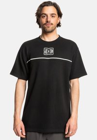 Black short-sleeve t-shirt made of cotton blend, featuring a white horizontal stripe and DC logo in the center. Loose fit with ribbed neckline.