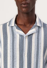 Camisa de botones a rayas que presenta tonos alternos de azul y blanco, confeccionada con un material texturizado, con un cuello suave y detalles de botones visibles.