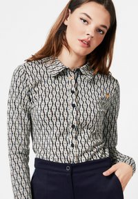 Button-up shirt with a geometric pattern in beige, navy, and light blue tones. Features a collar and long sleeves, made from a smooth fabric.