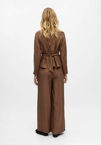 Femme avec de longs cheveux blonds portant un haut à manches longues marron noué à la taille et un pantalon large assorti, se tenant sur un fond blanc.