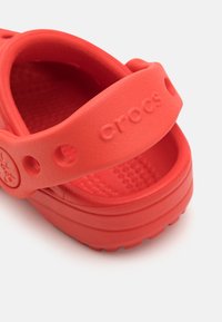 Punaiset kumiset puukengät, joissa on teksturoitu pohjallinen ja leveä hihna. Niissä on pyöreät ilmanvaihtoaukot, sileä pinta ja kohotettu pohja, joka parantaa pitoa.