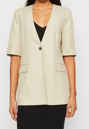 Beiger Blazer mit kurzen Ärmeln, Ein-Knopf-Verschluss und Pattentaschen, getragen über einem schwarzen Kleid mit V-Ausschnitt, dargestellt von den Schultern bis zur Hüfte.