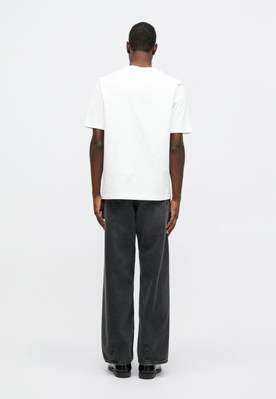 Muž stojící zády k fotoaparátu, oblečený v jednoduchém bílém tričku, volných černých džínách a černých botách na neutrálním pozadí.