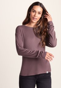 Long-sleeve mauve top made of ribbed fabric. Features a boat neckline and fitted cuffs. Paired with dark jeans and a visible brand tag.