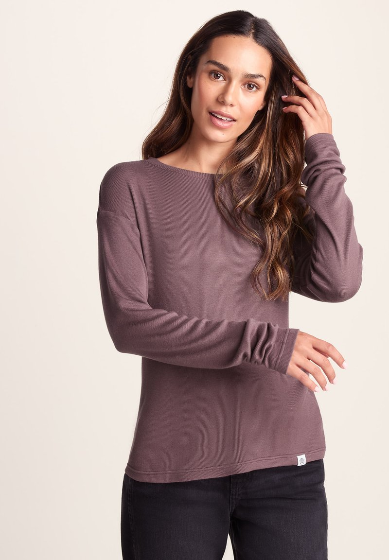 Long-sleeve mauve top made of ribbed fabric. Features a boat neckline and fitted cuffs. Paired with dark jeans and a visible brand tag.