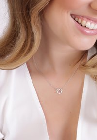 Zilveren ketting met een hartvormige hanger met daarin een oneindigheidssymbool, versierd met kleine rhinestones, gedragen tegen een witte blouse.