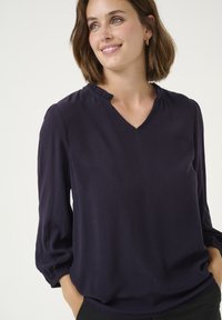 Navy blue blouse with a V-neck and ruffled neckline. Features long, slightly puffed sleeves and a textured fabric.