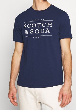 Mężczyzna ubrany w granatowy T-shirt z białym napisem „Amsterdam Scotch & Soda Blauw xxx” i jasne spodnie, stojący na białym tle.