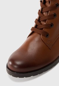 Bota de cuero marrón hasta el tobillo con acabado texturizado, punta redonda y ojetes de bronce. Presenta cordones marrones y una suela de goma negra.