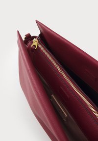 Burgunderlæder håndtaske med en tekstureret finish, der har en guldfarvet lynlås og to rum, som fremhæver et minimalistisk design.