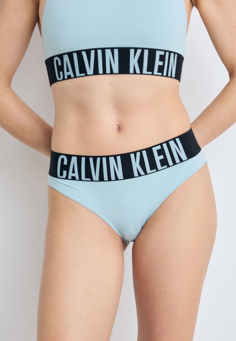 Lichtblauwe sportbh en bijpassende slip met een zwarte tailleband waarop "CALVIN KLEIN" in het wit staat, van gladde stof, minimalistisch ontwerp.