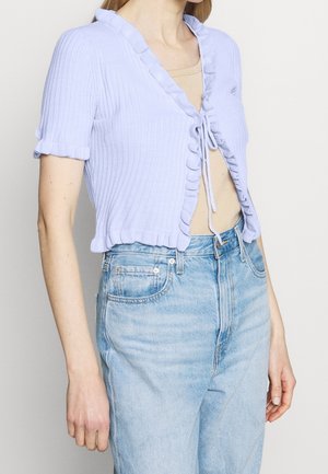 Žena ve světle modrém svetříku s krátkými rukávy s volánky, který je vepředu zavázaný, přes béžové tričko a ve světle modrých džínech s vysokým pasem.