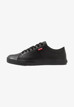Sapatilhas pretas feitas de material sintético texturizado, com um design de cano baixo, bico redondo, sola plana de borracha e logotipo vermelho da Levi's na lateral.