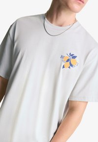 Světle šedé bavlněné tričko s krátkým rukávem, na levé části hrudi s grafikou modrožlutého citronu a textem.