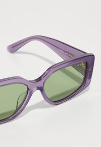 Μωβ πλαστικά γυαλιά ηλίου με πράσινους φακούς, γωνιακό σχέδιο, ευρείες γέφυρες και επωνυμία στο πλάι. Ελαφριά με λεία υφή.