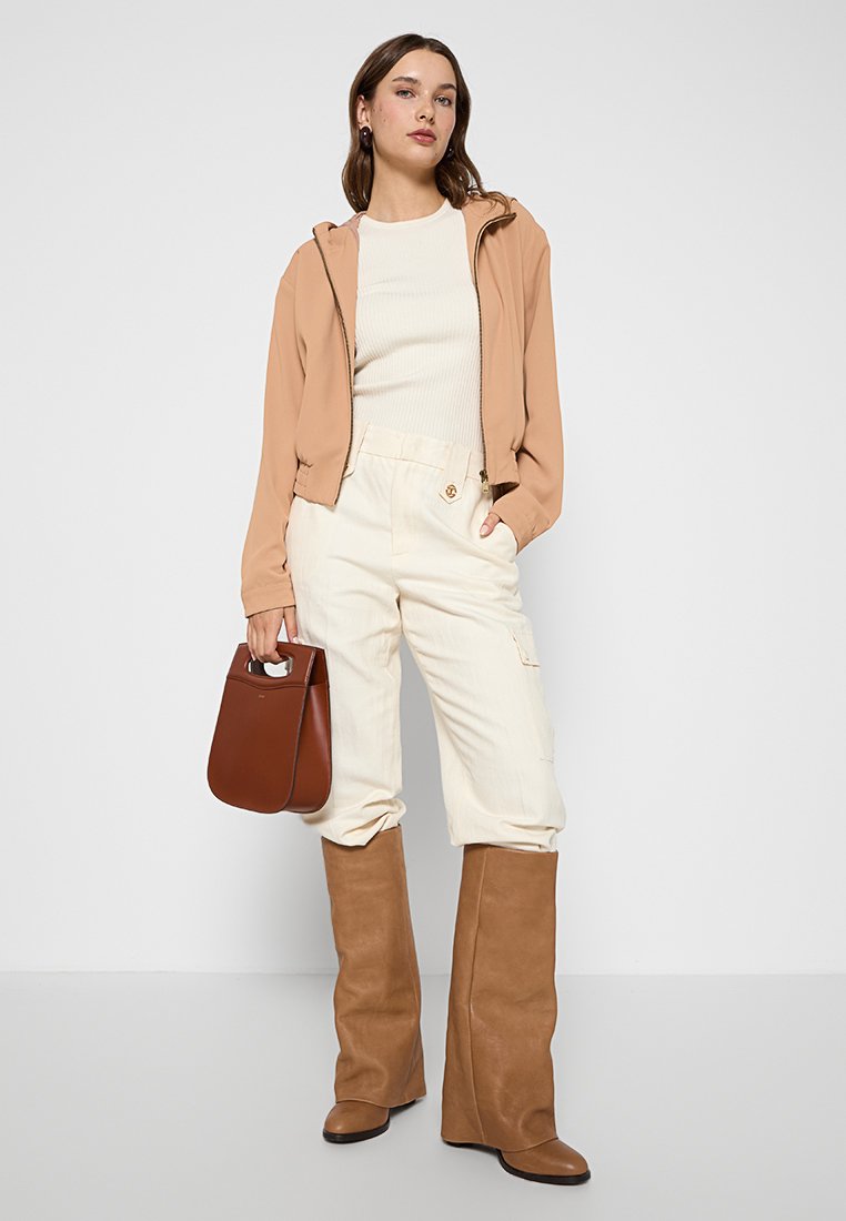 Chaqueta ligera beige con cremallera, top crema de canalé, pantalones blancos estilo cargo, botas marrones hasta la rodilla y un bolso marrón.