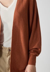 Rustfarvet strikket cardigan med løse ærmer, åben front og struktureret stof. Båret over en hvid ribbet V-udskæring top og khaki bukser.