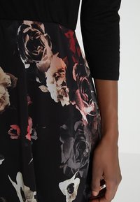 Schwarzes Kleid mit Dreiviertelärmeln; florales Muster in den Farben Rot, Weiß und Grau auf dem Rock; glatter Stofftexture.