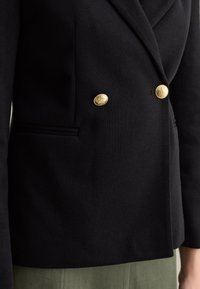 Blazer negro de corte doble, hecho de tejido texturizado, con botones dorados y dos bolsillos laterales. Cuello afilado y ajuste entallado.