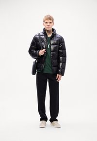 Zwarte pufferjack met een glanzende afwerking, ritsluiting, gecombineerd met een groene sweatshirt, zwarte broek en lichte sneakers, met een zwarte tas.