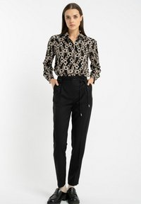 Kobieta stoi z rękami w kieszeniach, ubrana w bluzkę z czarnym wzorem łańcucha, wysokie czarne spodnie z paskiem i czarne mokasyny w jasnych skarpetkach.