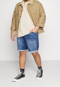 Beige corduroy takki vaalean beige t-paidan päällä, yhdistettynä sinisiin farkkushortseihin ja mustiin Converse-kengiin, shortseissa käännetyt hihansuut.