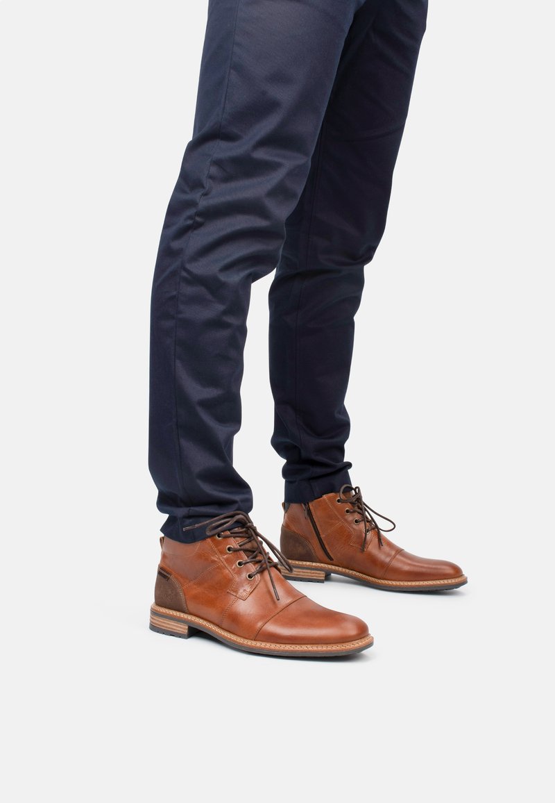 Botas de cuero marrón hasta el tobillo con un acabado suave, punta redonda, costuras decorativas y cordones marrones, combinadas con pantalones oscuros.