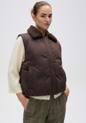 Donna che indossa un gilet imbottito marrone con colletto peloso, maglione bianco e pantaloni a quadri, in piedi con le mani nelle tasche davanti a uno sfondo semplice.