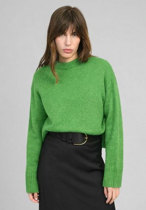 Femme aux cheveux bruns mi-longs portant un pull vert vif et une jupe noire avec une ceinture noire, debout devant un fond gris.
