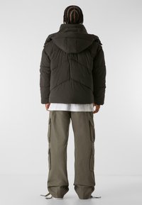 Puffjacke in Dunkelgrau mit Kapuze, mit horizontalen Nähten und lockerer Passform. Kombiniert mit olivgrünen Cargo-Hosen mit Seitentaschen.
