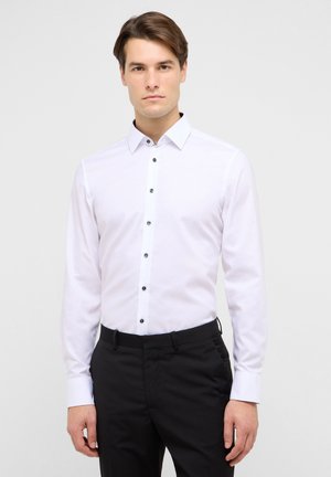 Camicia bianca a maniche lunghe con collo classico e bottoni neri, abbinata a pantaloni neri. Tessuto liscio con vestibilità su misura.