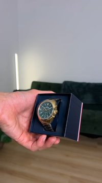 Ρολόι σε χρυσό τόνο με σκοτεινό πράσινο καντράν, μεταλλικό λουράκι σε ασημί τόνο και πολλαπλά καντράν, τοποθετημένο σε μαύρο κουτί παρουσίασης.