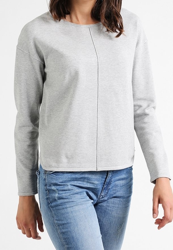 Femme portant une chemise à manches longues gris clair avec une couture centrale, associée à un jean bleu, sur un fond blanc uni.