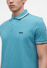 Homme portant un polo bleu clair avec un col et des poignets rayés noir et blanc, ainsi que le logo "BOSS" sur la poitrine gauche.