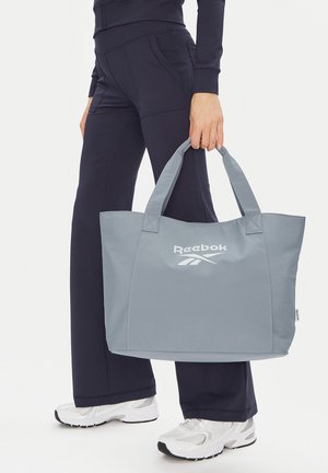 Persona con pantalones deportivos negros y zapatillas blancas sosteniendo una gran bolsa tote gris de Reebok por las asas.