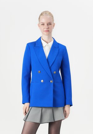 Blå dobbeltbrystet blazer med gullknapper, nøkkelklapper og strukturerte skuldre, kombinert med en hvit skjorte med krage og grå plisserte skjørt.