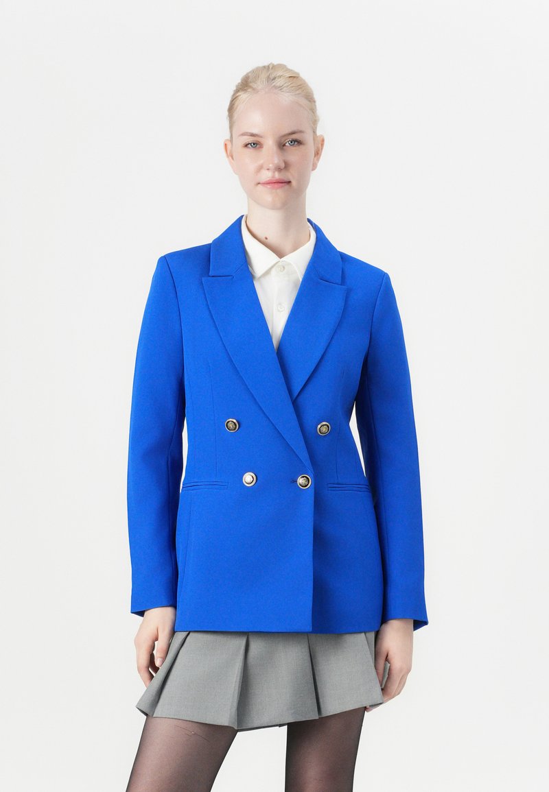 Modrý dvouřadý blazer se zlatými knoflíky, klopami a strukturovanými rameny, v kombinaci s bílou košilí s límcem a šedou skládací sukní.
